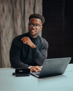 Un homme joyeux avec des lunettes travaillant sur un ordinateur portable à une table à l'intérieur, dégageant une ambiance d'espace de travail professionnelle et moderne.