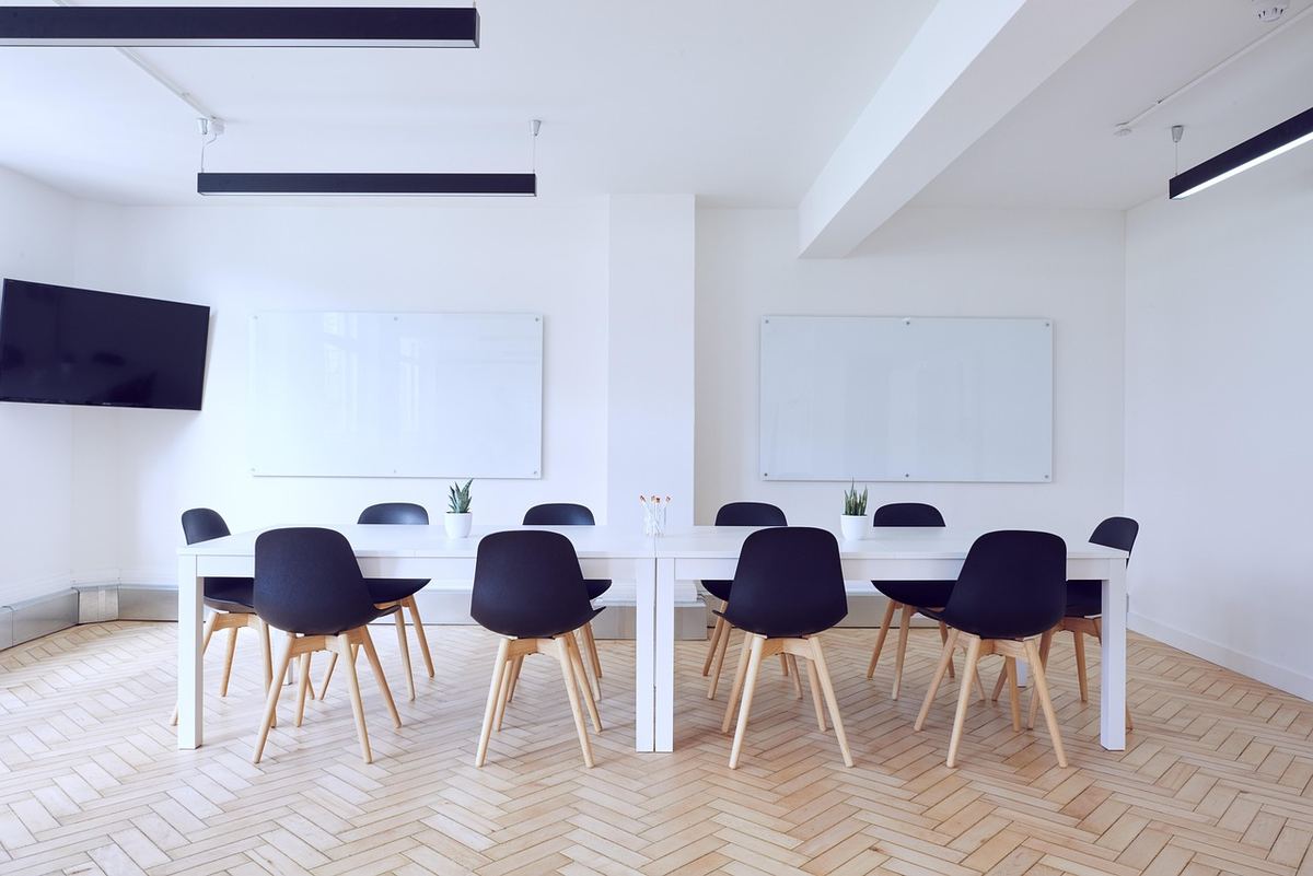 Salle claire avec table longue, chaises noires, tableau blanc et TV murale